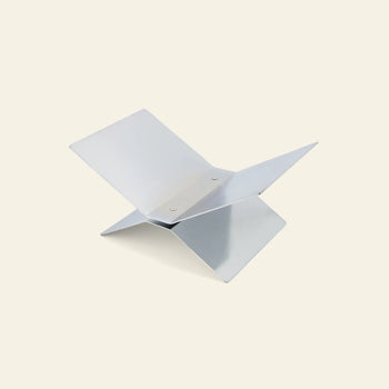 The baebsy Atlas Bookstand by baebsy features a minimalist, modern design with two interlocking flat panels forming an X shape, creating a levitation effect. It is shown against a plain light background.
