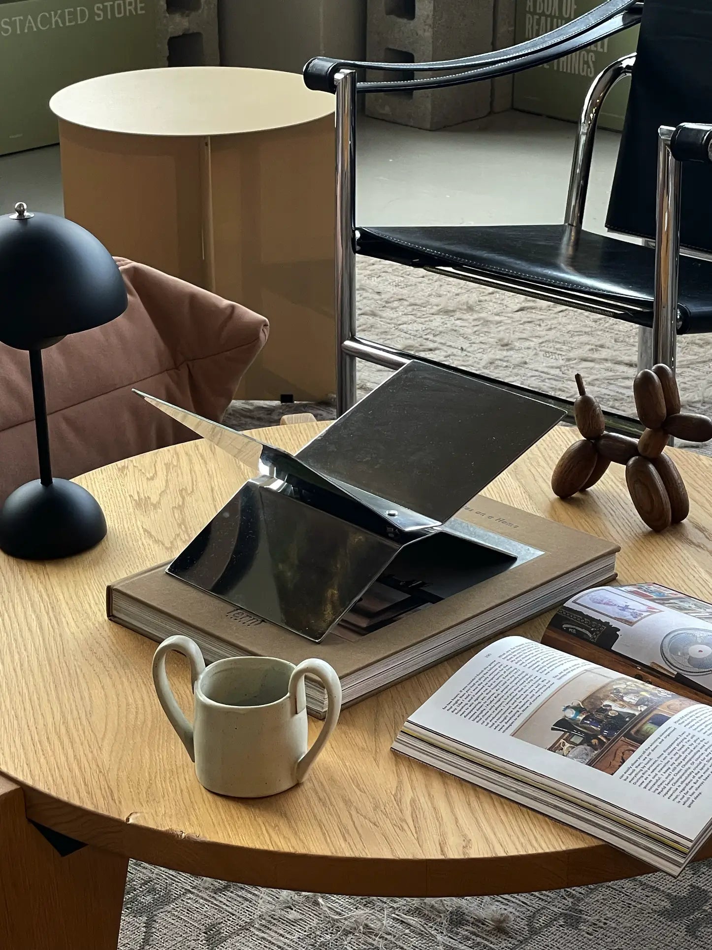 A modern wooden coffee table with an open book on a baebsy Atlas Bookstand, a beige mug, black lamp, metallic laptop stand with a closed laptop, small sculpture, and a chair in the background.