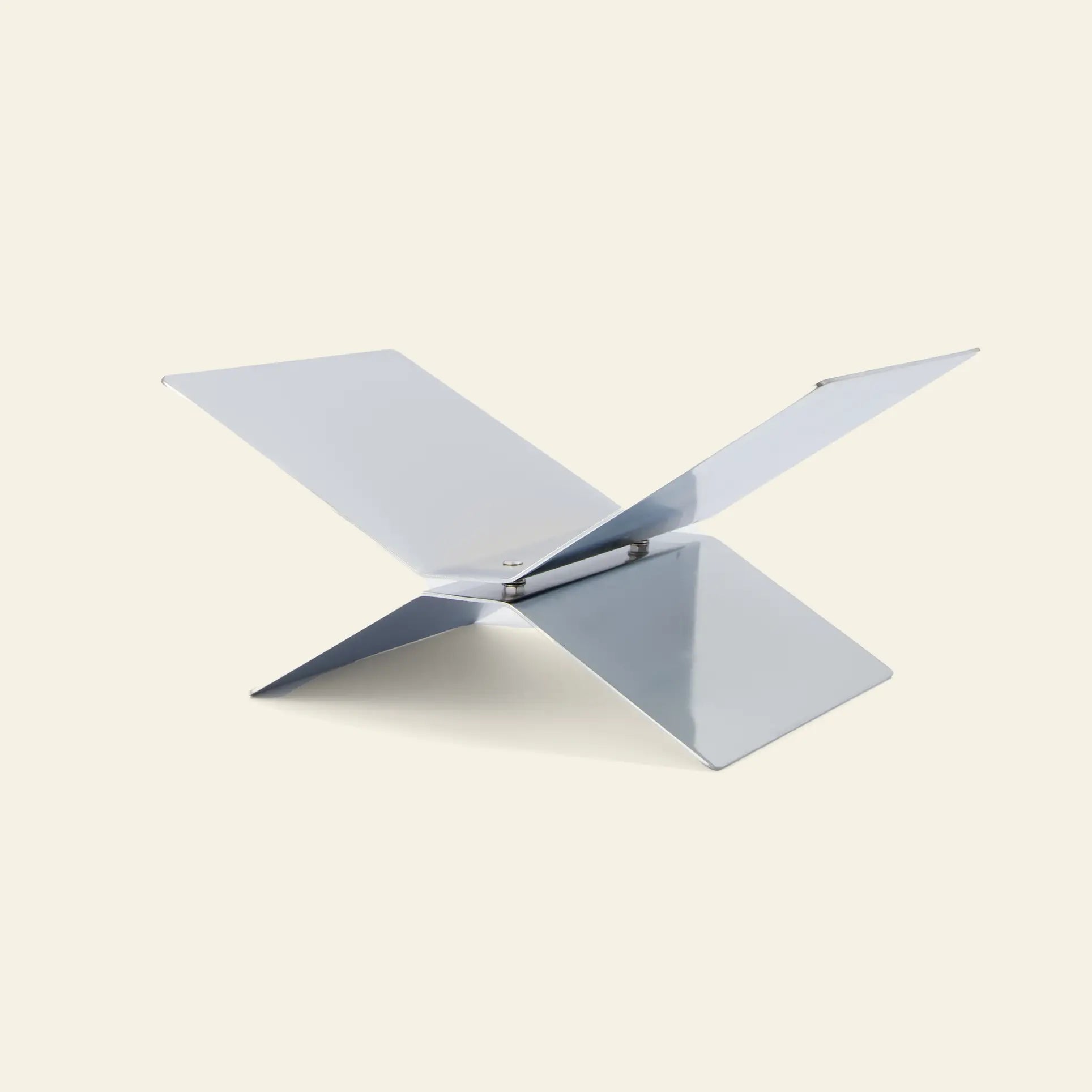 The baebsy Atlas Bookstand by baebsy is a minimalist, metallic bookstand with an X-shaped design formed by four flat intersecting panels, displayed on a light beige background.