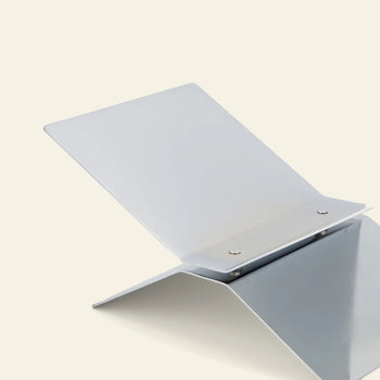 The baebsy Atlas Bookstand, a minimalist silver metal stand with a sleek, angled design by baebsy, is displayed against a light beige background.