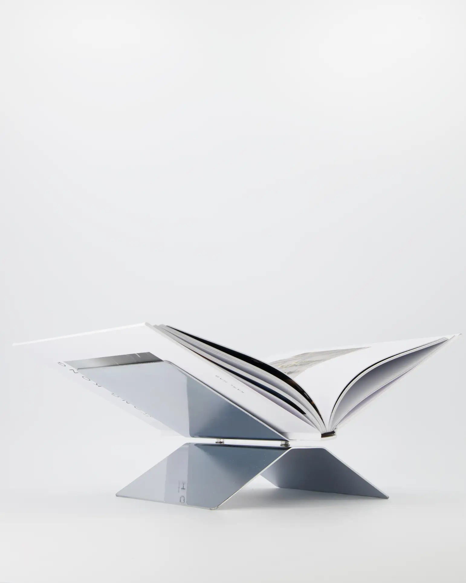 The baebsy Atlas Bookstand by baebsy features a minimalist X-shaped design with two intersecting panels, creating a levitating effect. Its sleek assembly stands out on a stark white background, making it ideal for showcasing open books.