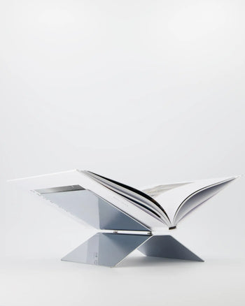 The baebsy Atlas Bookstand by baebsy features a minimalist X-shaped design with two intersecting panels, creating a levitating effect. Its sleek assembly stands out on a stark white background, making it ideal for showcasing open books.