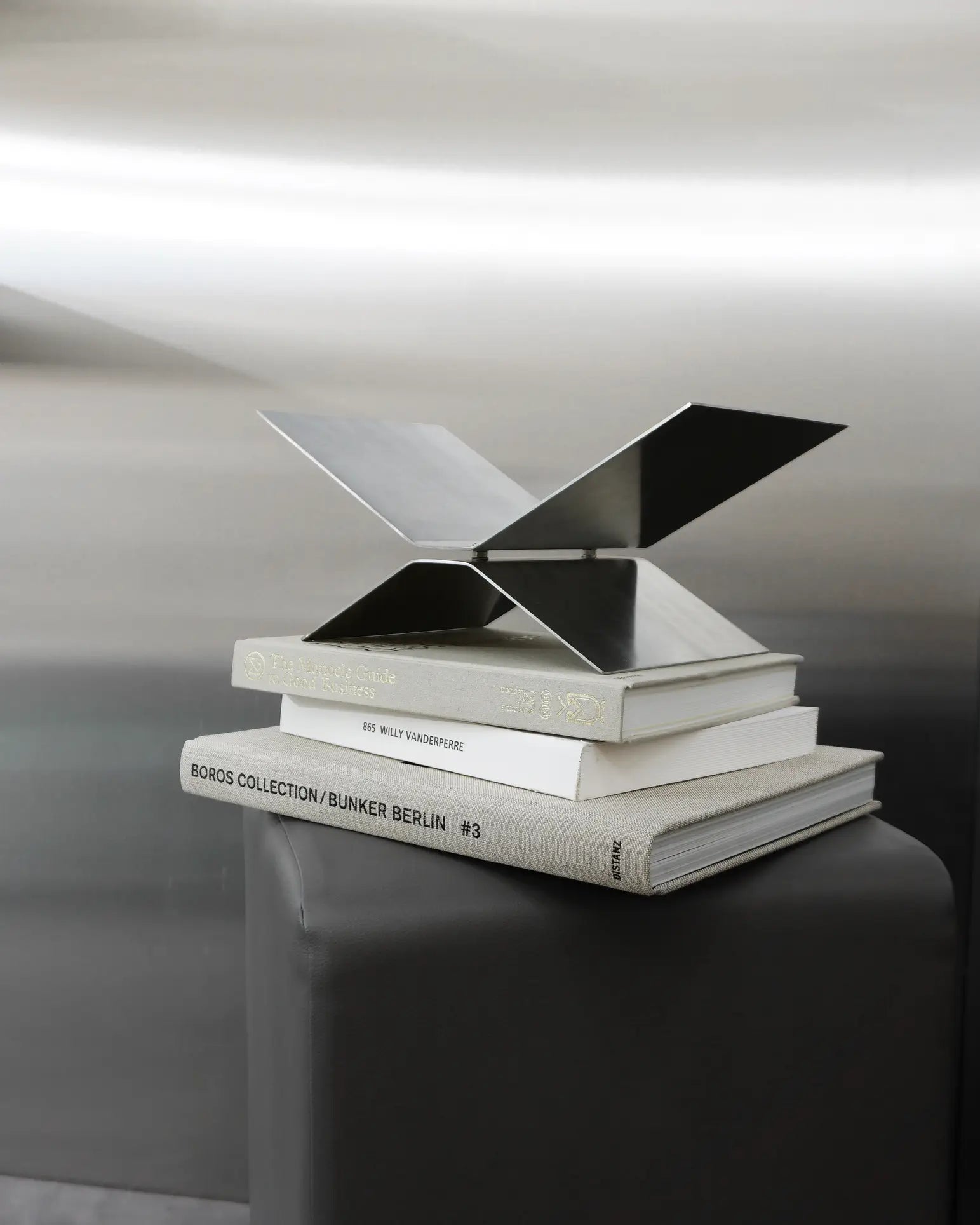 The baebsy Atlas Bookstand by baebsy, with its modern X-shaped design and levitation effect, is displayed on a stack of three large art books atop a gray pedestal, standing out against a metallic background.