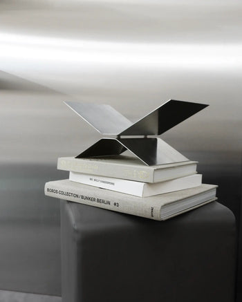 The baebsy Atlas Bookstand by baebsy, with its modern X-shaped design and levitation effect, is displayed on a stack of three large art books atop a gray pedestal, standing out against a metallic background.