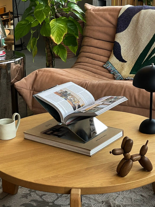 A modern living room with a tan lounge chair, leafy plant, wooden coffee table holding a magazine on a metal stand, ceramic mug, and an &Tradition Setago JH27 Portable Table Lamp beside a wooden balloon dog sculpture.