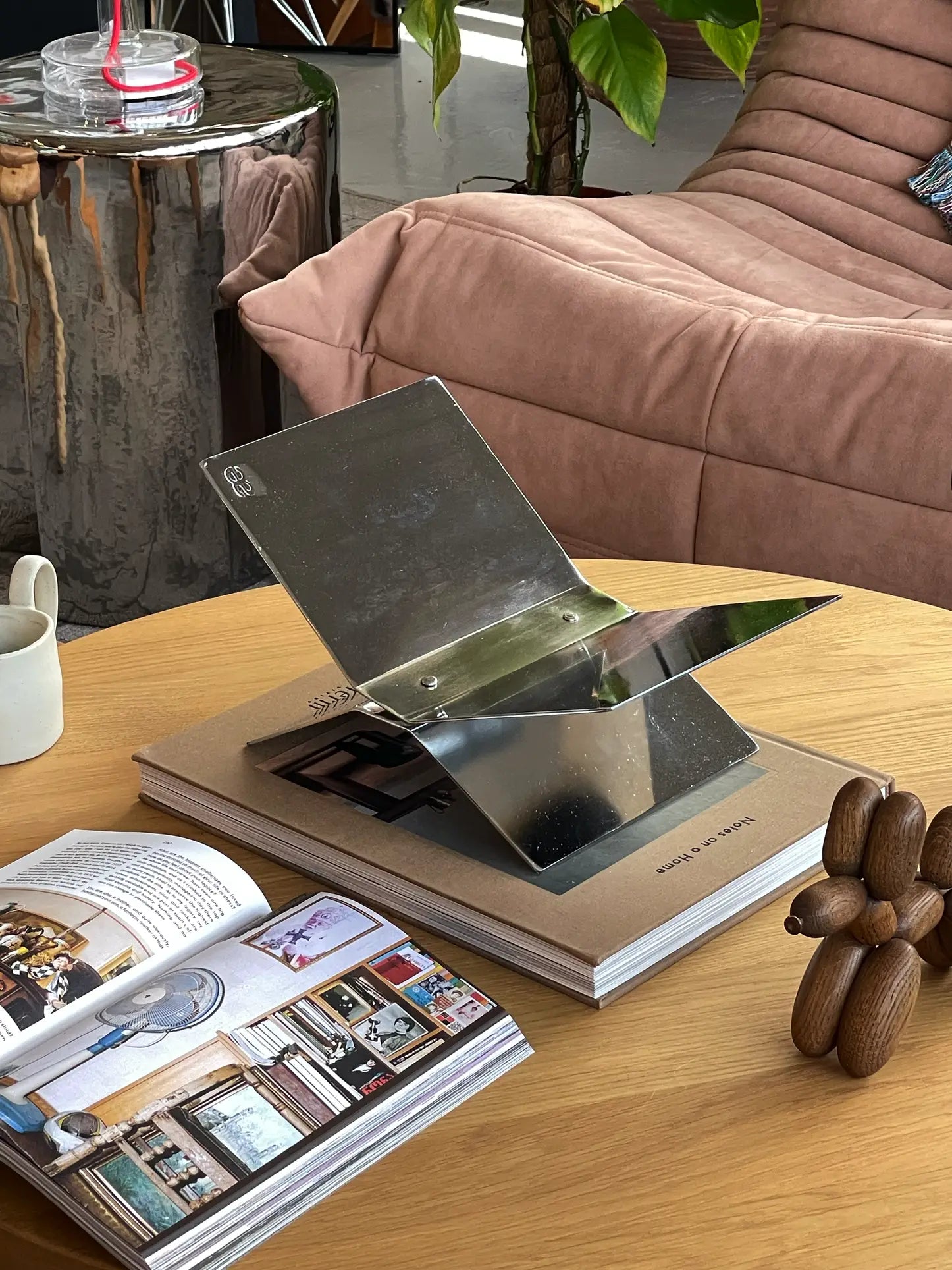 A modern coffee table with an open magazine, a sculptural wooden object, the baebsy Atlas Bookstand displaying its levitation design, a small white cup, and a beige couch near a green plant in the background.