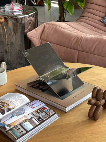 A modern coffee table with an open magazine, a sculptural wooden object, the baebsy Atlas Bookstand displaying its levitation design, a small white cup, and a beige couch near a green plant in the background.