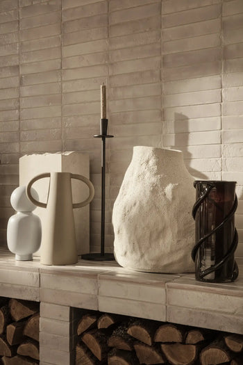 The ferm LIVING Anse Vase by ferm LIVING sits on a white tiled mantel above stacked firewood, accompanied by a sculptural object and tall candlestick, all set against a textured, light-colored tiled wall.