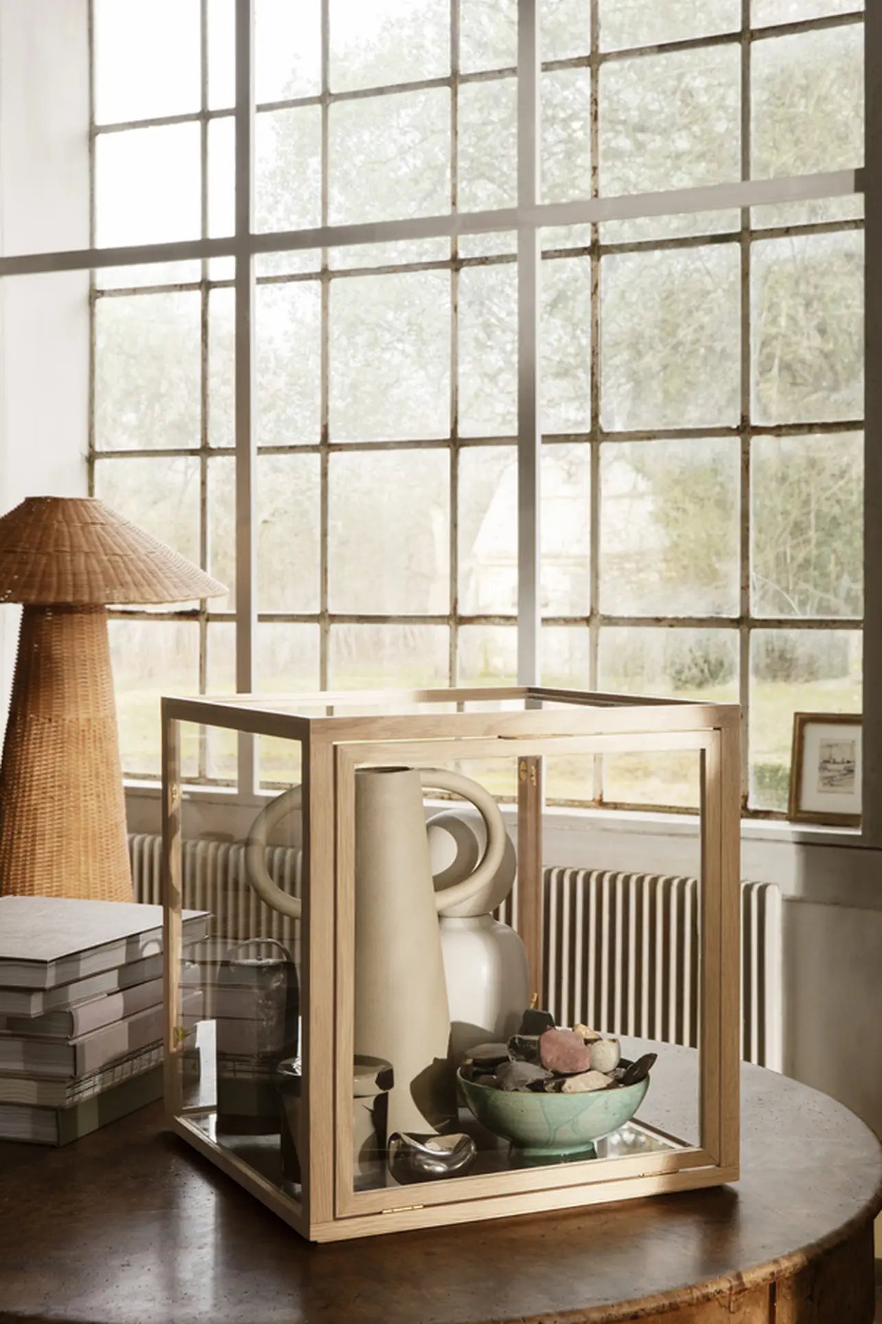 A wooden and glass display case rests on a round table, featuring the ferm LIVING Anse Vase by ferm LIVING alongside a bowl of decor. In the background are stacked books, a wicker lamp, and a large grid window with garden views.