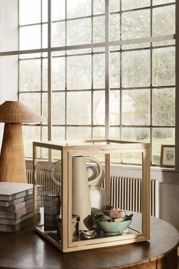 A wooden and glass display case rests on a round table, featuring the ferm LIVING Anse Vase by ferm LIVING alongside a bowl of decor. In the background are stacked books, a wicker lamp, and a large grid window with garden views.
