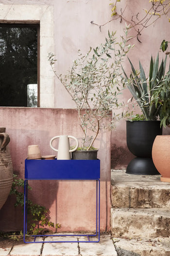 A ferm LIVING Anse Vase sits alongside a potted olive tree and beige watering can against a weathered pink wall, with rustic clay pots and lush greenery on stone steps; a window reflects the outdoors.