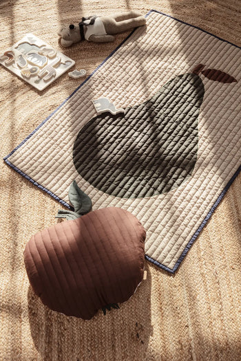 A ferm LIVING Apple Quilted Cushion rests on a woven rug beside a quilted cotton play mat, plush toy, and wooden animal tray in a childs room, with sunlight casting gentle shadows across the scene.