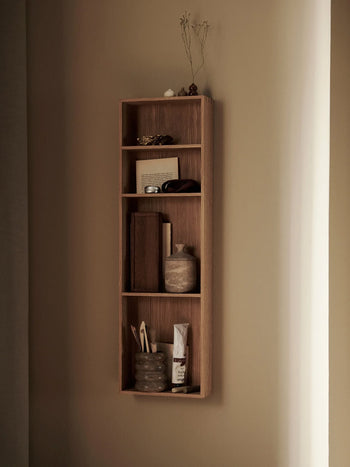 A wooden wall-mounted shelf displays decorative objects, including a ferm LIVING Bendum Cup, a framed photo, books, dried flowers, and a bowl—all elegantly arranged against a beige wall with soft lighting.