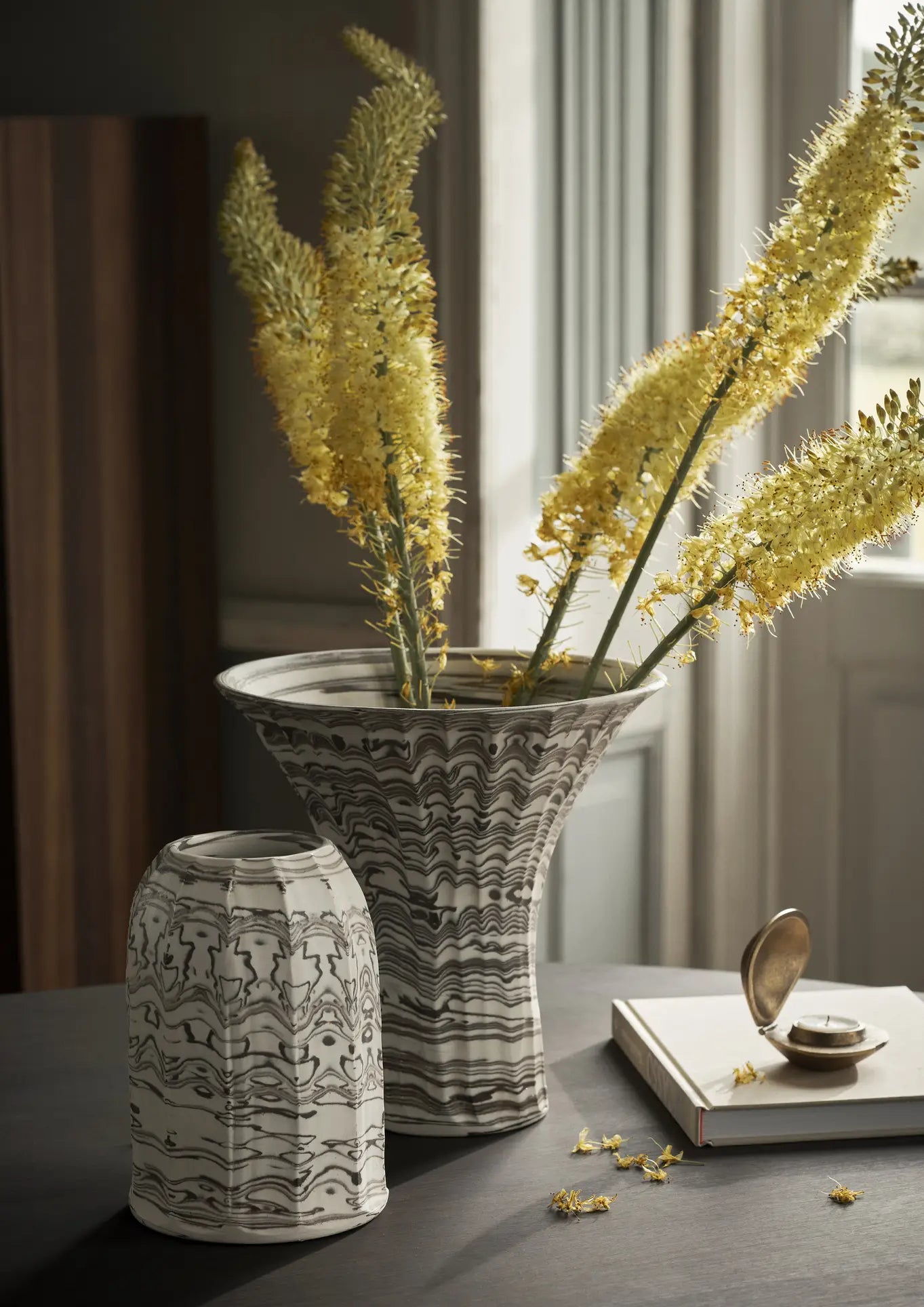 The ferm LIVING Blend Vase, Large, features a marbled black and white design with tall yellow flowers. An upside-down matching piece, pocket watch, and small yellow flowers sit on a dark table by a softly lit window.