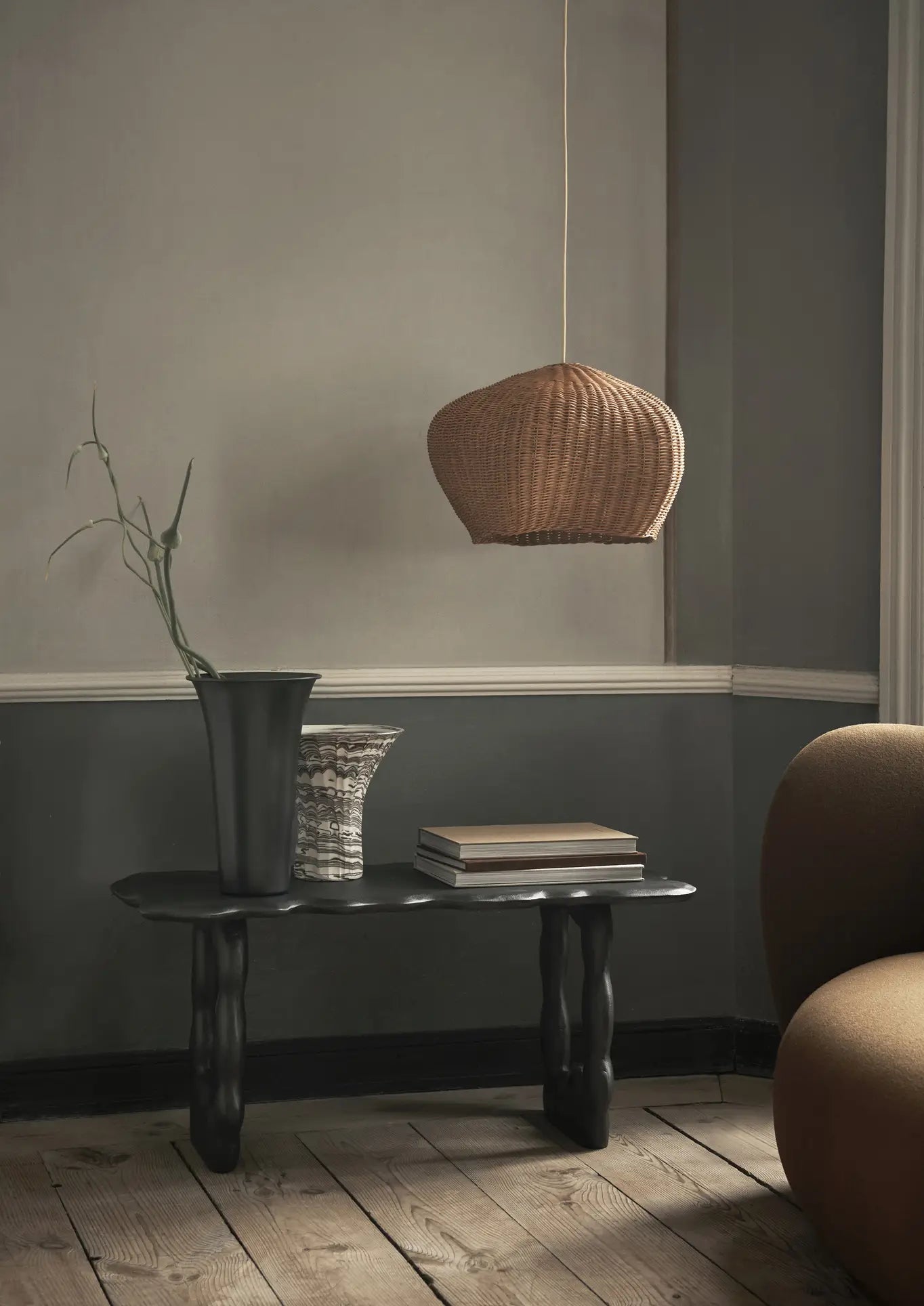 A minimalistic room with earthy tones features a dark sculpted table holding the ferm LIVING Blend Vase, Large by ferm LIVING, a stack of books, and dried foliage. A woven pendant lamp hangs above. Gray walls, a brown armchair, and wooden floors complete the look.