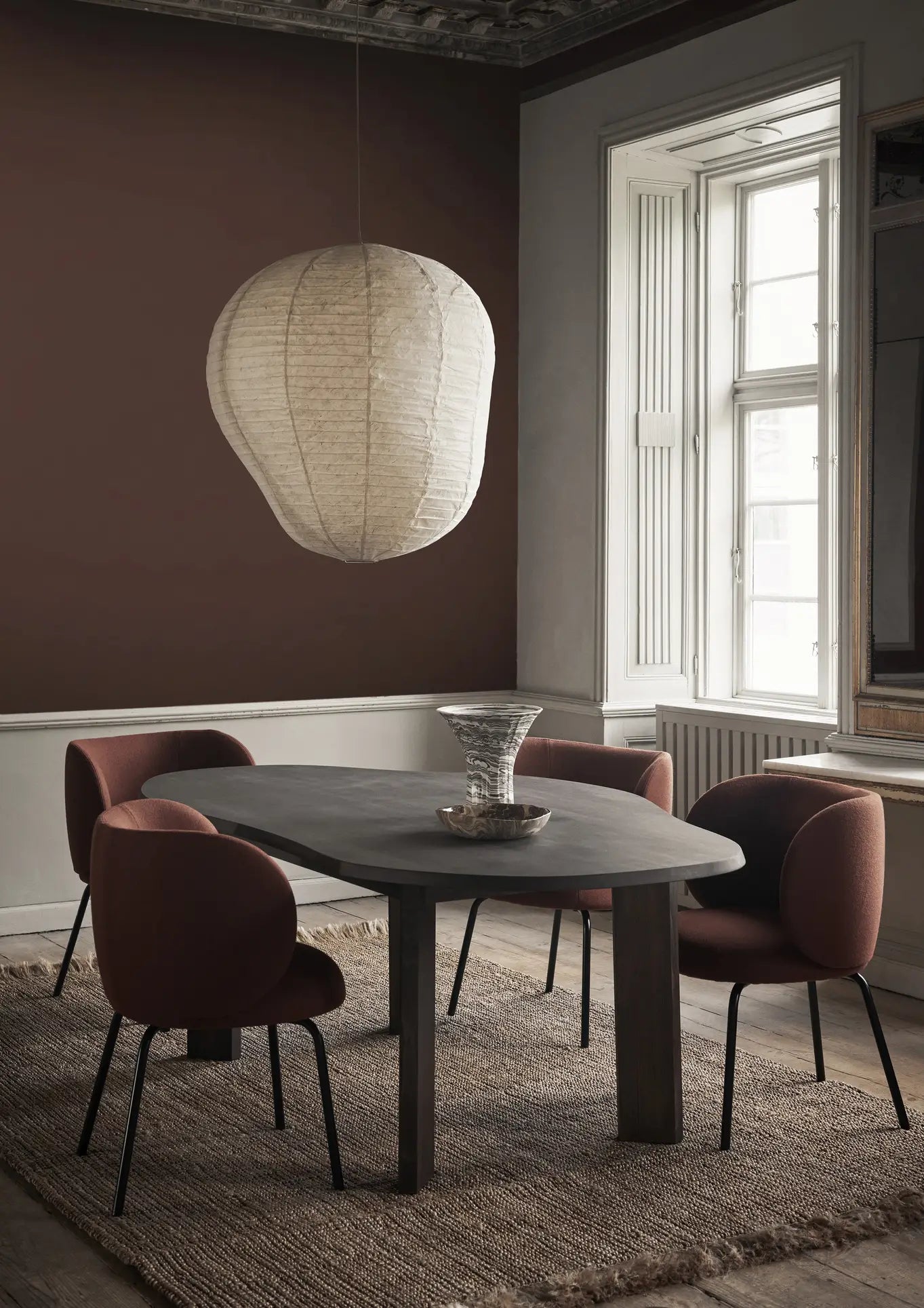 A modern dining room with a dark oval table, four reddish-brown upholstered chairs, a large paper lantern pendant light, and the ferm LIVING Blend Vase, Large by ferm LIVING on a textured rug near a tall window with white shutters.