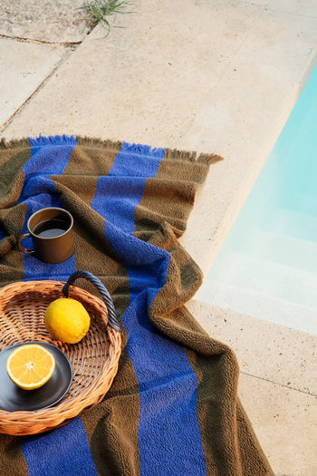 A striped blue and brown towel lies by the pool, with the ferm LIVING Blue Handle Tray from ferm LIVING holding a cup of coffee, a lemon, and a halved orange on a black plate, creating an inviting poolside scene.