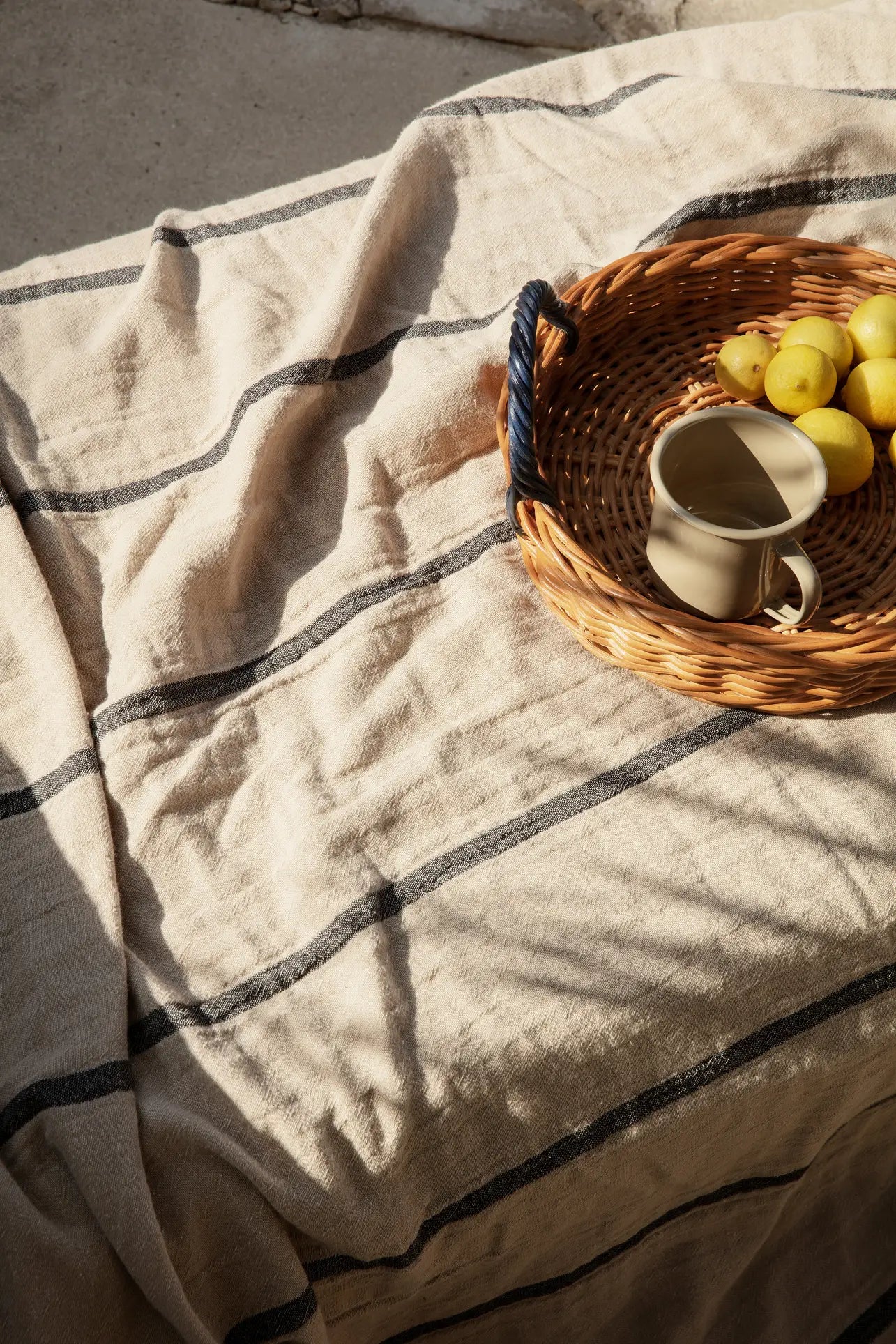 The ferm LIVING Blue Handle Tray by ferm LIVING, topped with yellow lemons and a ceramic mug, rests on a cream-striped cloth and makes a charming sunlit decorative accent.