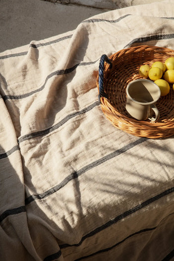 The ferm LIVING Blue Handle Tray by ferm LIVING, topped with yellow lemons and a ceramic mug, rests on a cream-striped cloth and makes a charming sunlit decorative accent.