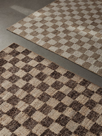 Two checkered rugs sit on a grey floor. The rug in front is the ferm LIVING Check Wool Jute Rug (200x300) with a darker brown and tan checkerboard design, while the one in back displays a light beige and cream check pattern.