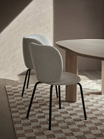 Two modern, light gray upholstered chairs with black legs sit beside a light wood table on a ferm LIVING Check Wool Jute Rug (200x300) by ferm LIVING in a softly lit, minimalist room.