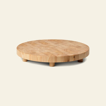 The ferm LIVING Chess Cutting Board, Round Large, is crafted from FSC-certified larch wood with a distinctive grain and features short, sturdy legs. Shown here against a plain light background.