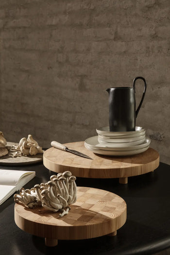 The ferm LIVING Chess Cutting Board, Round Large, made from FSC-certified larch wood, serves mushrooms alongside stacked white plates and a black pitcher, creating a minimalist rustic look against a textured gray wall.