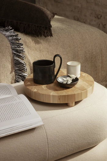 A round ferm LIVING Chess Cutting Board (Large) serves as a tray on a cushioned surface, holding a black mug, white candle, and candy dish. An open book and beige throw blanket rest nearby on the beige couch.