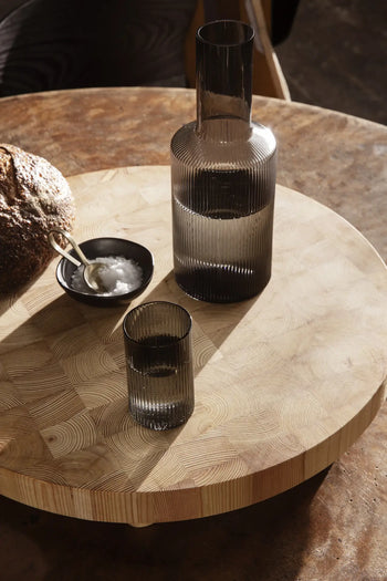 A glass carafe and matching glass sit on the ferm LIVING Chess Cutting Board, Round Large by ferm LIVING, with a bowl of salt, a small spoon, and rustic bread. Warm sunlight falls across the FSC-certified larch wood scene.