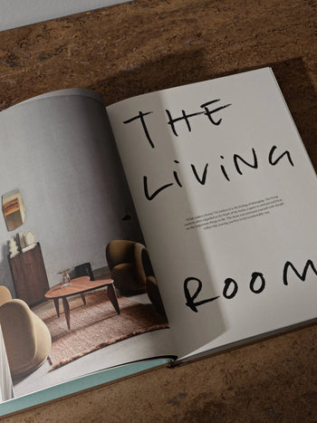 The ferm LIVING Coffee Table Book 2023 by ferm LIVING is open on a brown surface, featuring a modern living room photo on the left and large handwritten “THE LIVING ROOM” text over minimal text on the right.