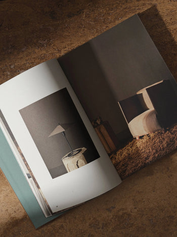 The ferm LIVING Coffee Table Book 2023 by ferm LIVING is open on a brown surface, featuring minimalist interior photos—a modern lamp on a stone table and a contemporary chair on a textured rug.