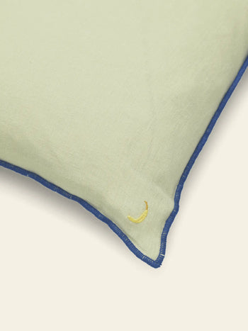 A close-up of the ferm LIVING Contrast Linen Cushion in light green, featuring blue contrast stitching and a small yellow crescent moon embroidered in one corner, set against an off-white background.