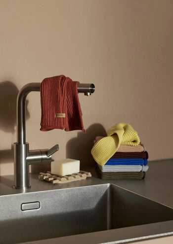 A modern kitchen sink with a stainless-steel faucet, a rust-colored ferm LIVING Day Cloth draped over it, a bar of soap on a wooden dish, and a stack of GOTS-certified ferm LIVING Day Cloths in various colors on the counter.