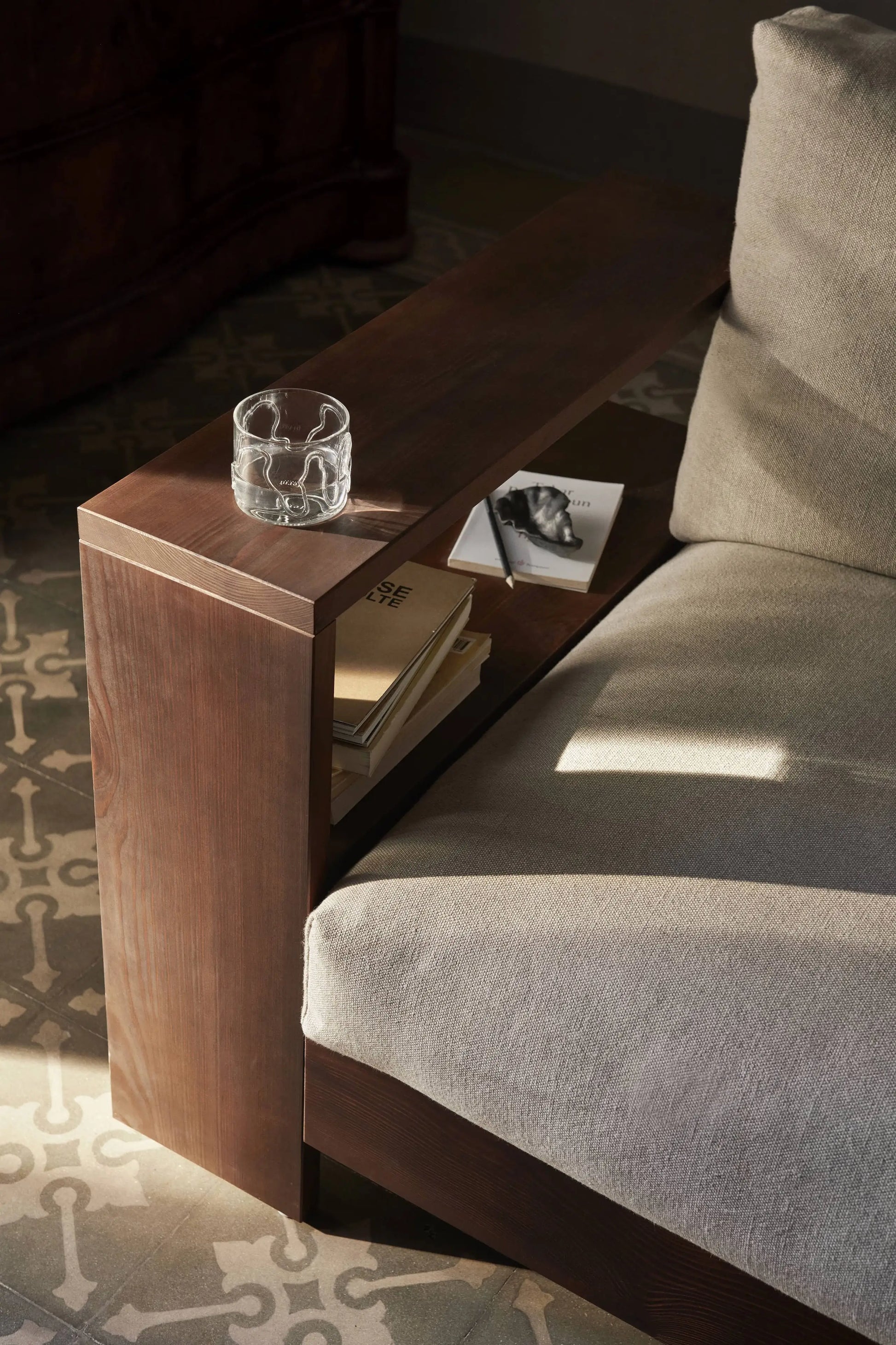 A close-up of a sofa with a wooden armrest featuring built-in shelves holding books and magazines. A ferm LIVING Doodle Glasses Low, Set of 2 glass of water sits on top, while the carpet in the background displays unique patterns.