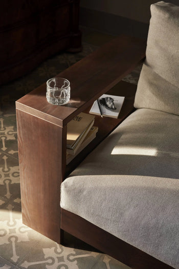 A close-up of a sofa with a wooden armrest featuring built-in shelves holding books and magazines. A ferm LIVING Doodle Glasses Low, Set of 2 glass of water sits on top, while the carpet in the background displays unique patterns.