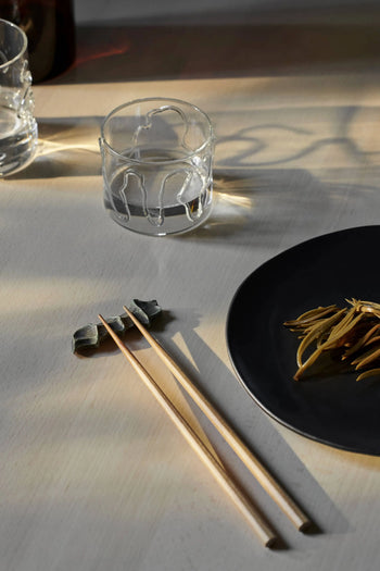 A ferm LIVING Doodle Glasses Low, Set of 2 by ferm LIVING with unique patterns, wooden chopsticks on a ceramic rest, and a black plate with food are elegantly arranged on a light table in natural sunlight.
