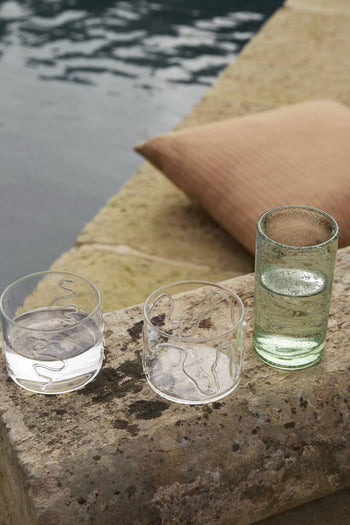 Two ferm LIVING Doodle Glasses Low sit on a stone ledge by the water—one filled with water, one empty—beside a tall glass. A beige cushion lies nearby. All glasses are crafted from borosilicate glass.