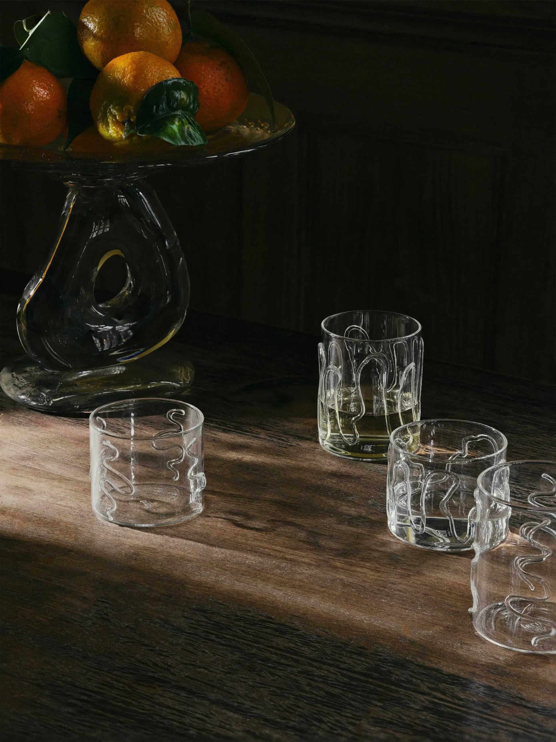 A wooden table with two sets of ferm LIVING Doodle Glasses Low—borosilicate glass tumblers with unique patterns, two filled and two empty—beside a glass fruit stand with oranges, sunlight casting shadows across the scene.