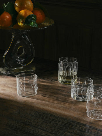 A wooden table with two sets of ferm LIVING Doodle Glasses Low—borosilicate glass tumblers with unique patterns, two filled and two empty—beside a glass fruit stand with oranges, sunlight casting shadows across the scene.