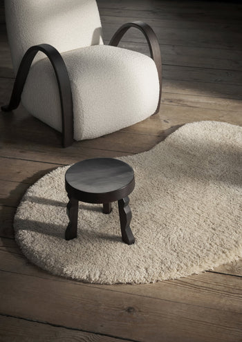 The ferm LIVING Faye Stool, a small round black accent, sits on a cream, irregular-shaped rug beside a modern white armchair with dark wooden arms atop wooden floorboards in a softly lit room.