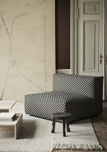 A modern interior showcases a checkered black-and-white lounge chair on a light rug, paired with the ferm LIVING Faye Stool and minimalist FSC-certified Mango wood tables, set against neutral walls and a large door in the background.