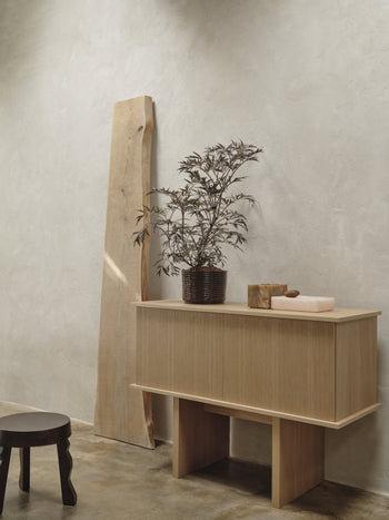 A minimalist interior features a light FSC-certified mango wood sideboard with a potted plant, two decor objects, the ferm LIVING Faye Stool as a dark accent piece, and a tall wooden plank leaning against a textured light wall.