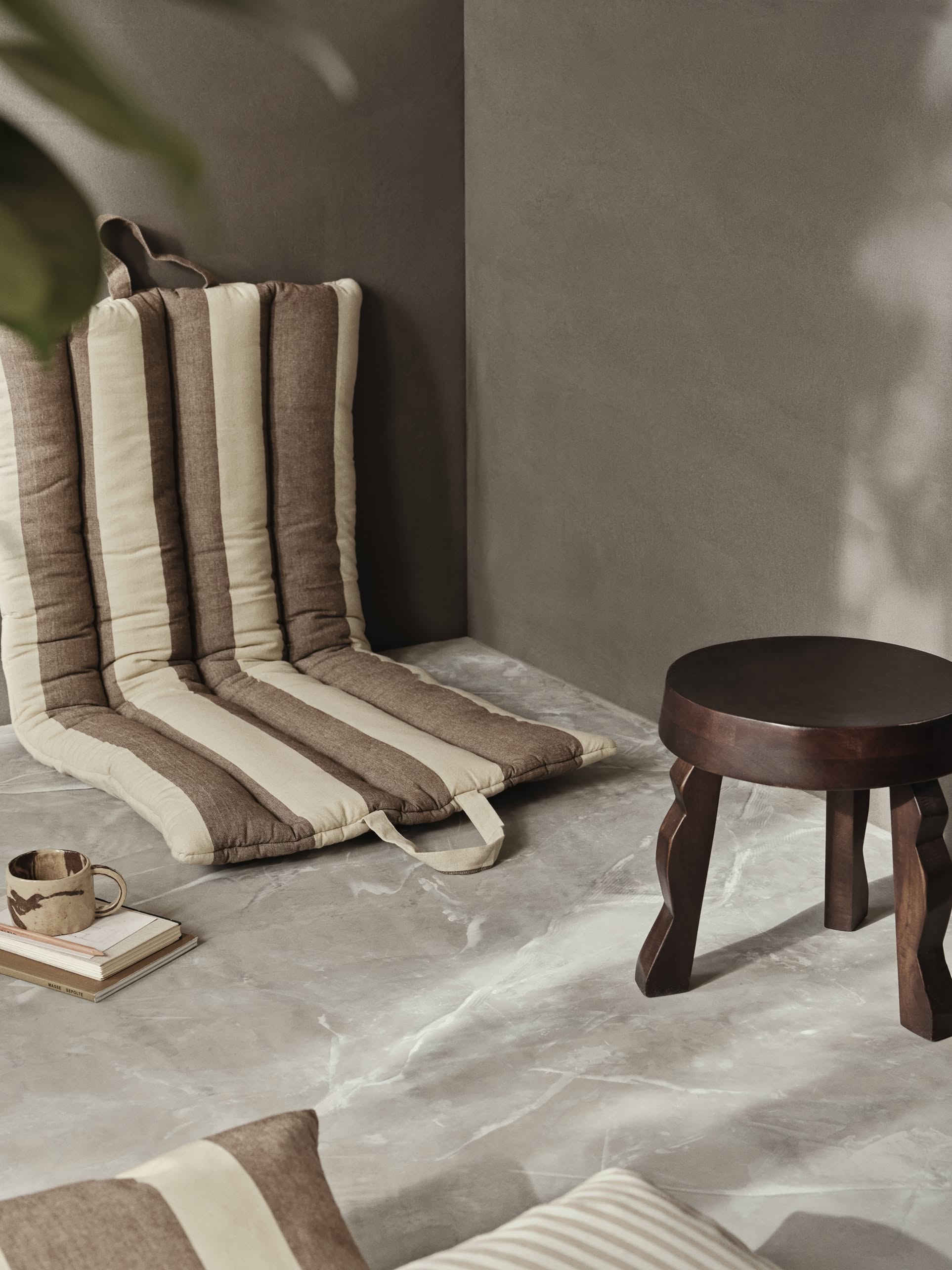 The ferm LIVING Faye Stool, crafted from dark FSC-certified mango wood, sits beside a beige and brown striped floor cushion against a gray wall, with a cup, book, and matching pillow arranged on the marble floor.