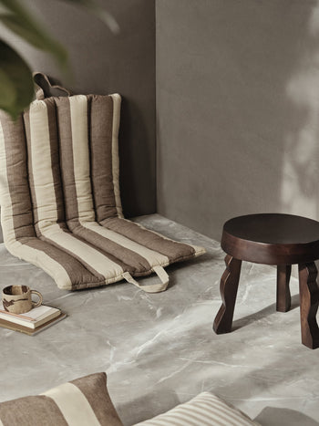 The ferm LIVING Faye Stool, crafted from dark FSC-certified mango wood, sits beside a beige and brown striped floor cushion against a gray wall, with a cup, book, and matching pillow arranged on the marble floor.