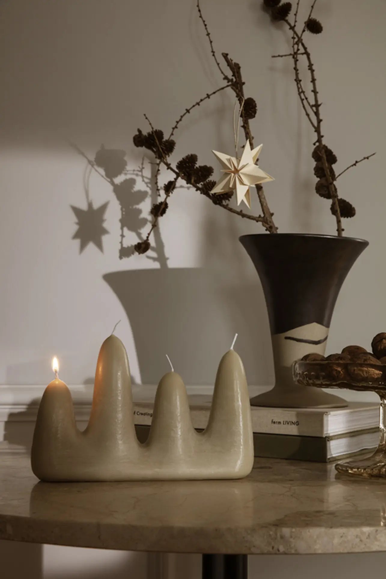 A wavy beige candle with four peaks, one lit, sits on a round table. Behind it, bare branches and a star ornament fill the ferm LIVING Flores Vase by ferm LIVING. Books are stacked on the right side of the table.