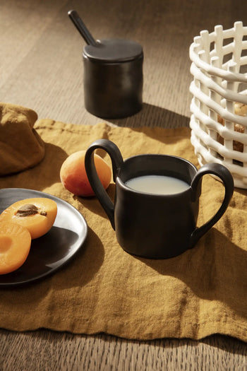 The ferm LIVING Flow Mug Mini in black, designed for children with two handles and filled with milk, sits on a mustard yellow cloth beside modern tableware—a plate of apricots, a black jar with spoon, and a white woven basket on a wooden table.