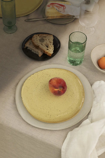 A table set with modern tableware: a yellow ferm LIVING Flow Plate, Medium holding a peach, a glass of water, bread on a dark semi-matte plate, pale napkin, and another plate with an egg—all on a beige tablecloth.