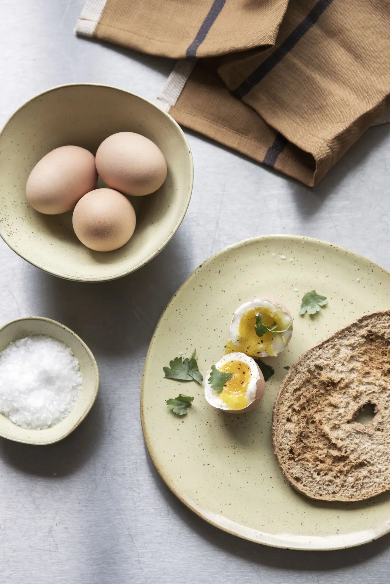 A halved soft-boiled egg with parsley, bread, and bowls of eggs and salt are served on the ferm LIVING Flow Plate, Medium by ferm LIVING, featuring a semi-matte glaze, set with a brown napkin on a gray surface.