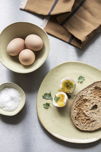 A halved soft-boiled egg with parsley, bread, and bowls of eggs and salt are served on the ferm LIVING Flow Plate, Medium by ferm LIVING, featuring a semi-matte glaze, set with a brown napkin on a gray surface.
