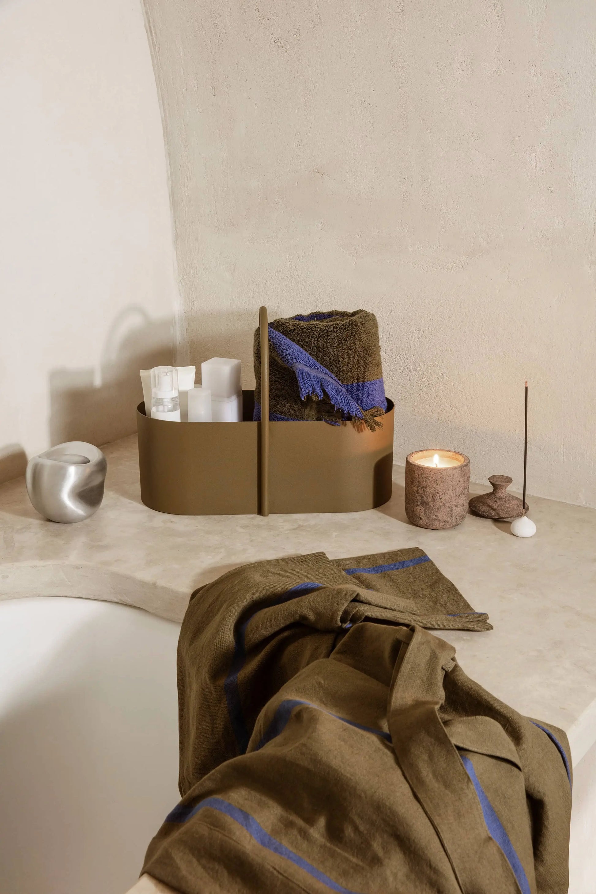 A bathroom scene features a beige counter with a brown caddy, toiletries, rolled towels, a lit candle, an incense holder, and the ferm LIVING Komo Mini Vases Set of 3. A blue-striped towel is draped over the bathtub’s edge.